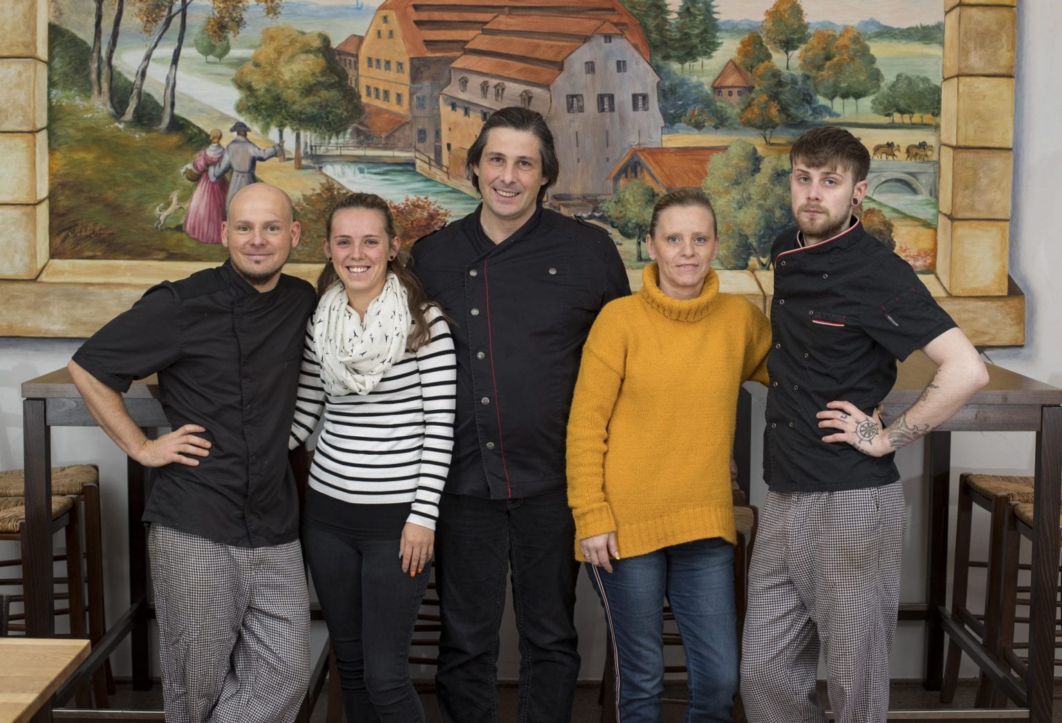 Team Dinner-Schmiede steht lächelnd in einem Raum, hinter ihnen hängt ein großes Gemälde einer rustikalen Landschaft an der Wand.