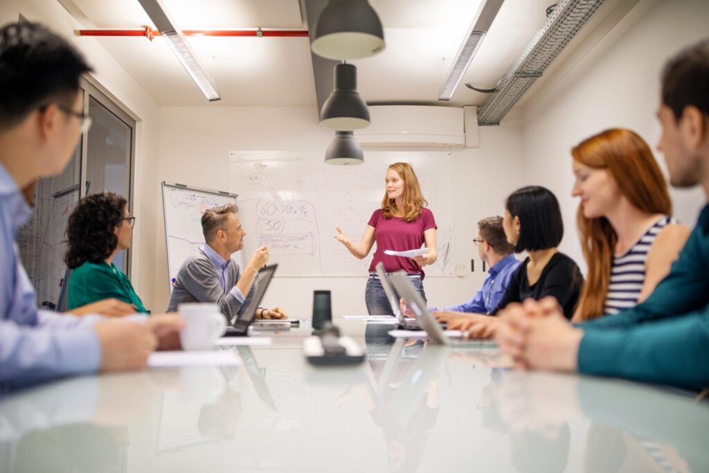 Frau präsentiert vor Gruppe mit Laptops, Whiteboard und Flipchart im Hintergrund.