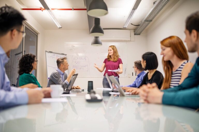 Frau präsentiert vor Gruppe mit Laptops, Whiteboard und Flipchart im Hintergrund.