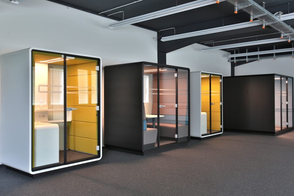 Mehrere Büroboxen in Weiß und Schwarz stehen im offenen, modernen Büro; kleine gelbe Telefonboxen und große Meetingboxen mit Glasfronten.