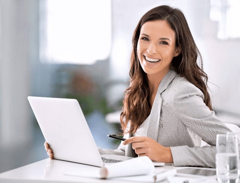 Frau am Schreibtisch mit Laptop, lächelnd, Stift in der Hand, Bürohintergrund.
