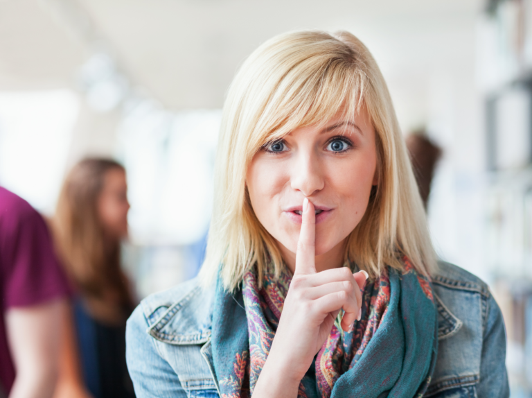 Frau mit Finger vor Lippen signalisiert Schweigen, unscharfer Hintergrund.