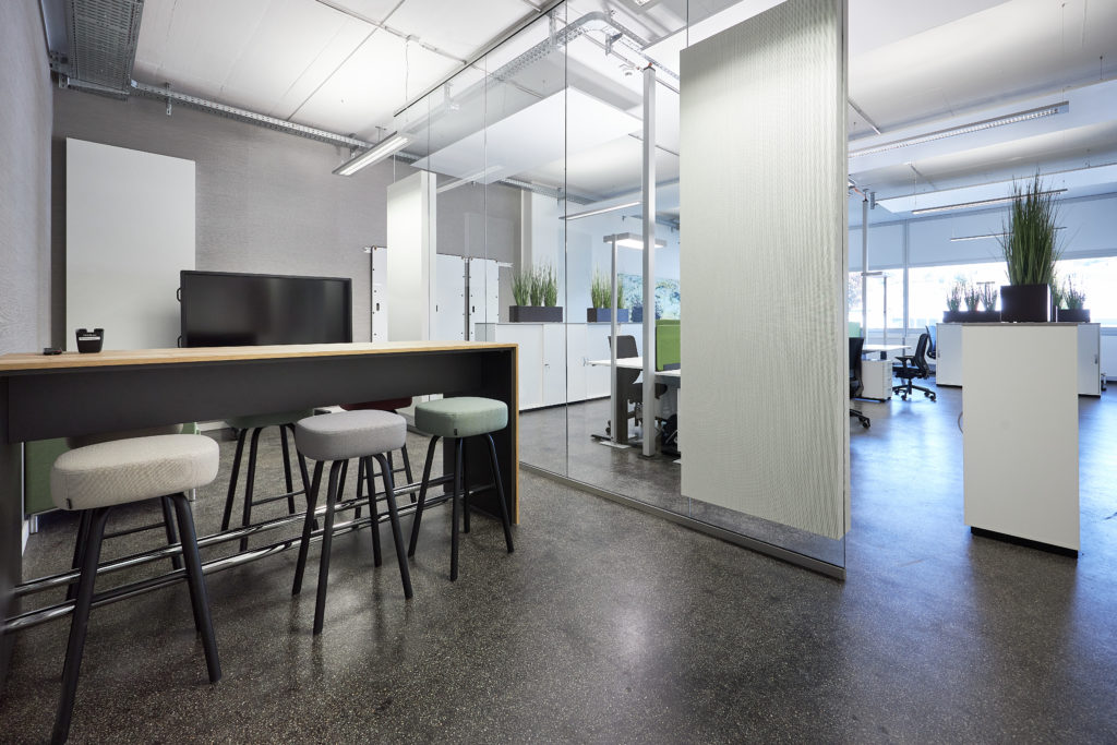 Hocker umgeben einen hohen Holztisch mit Monitor in modernem Büro mit Glaswänden und Pflanzen.