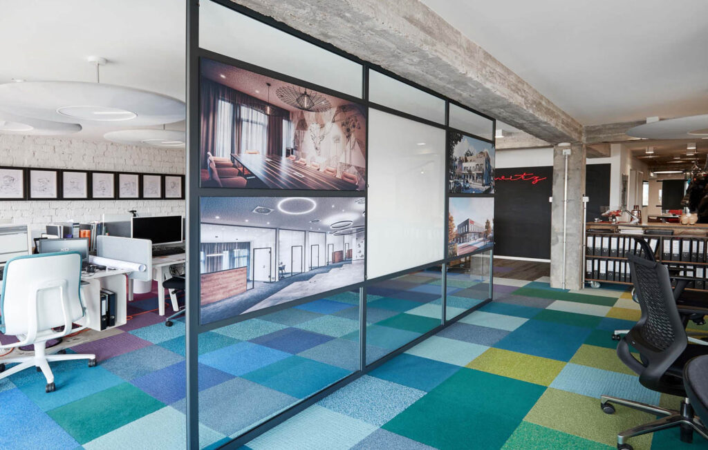 Glastrennwand mit Architekturmotiven trennt modernes Büro; bunte Teppichfliesen, Fotos an Wand.