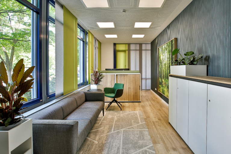 Bürolobby mit grauem Sofa, grünem Stuhl, Empfangstresen und Wald-Wandgemälde.