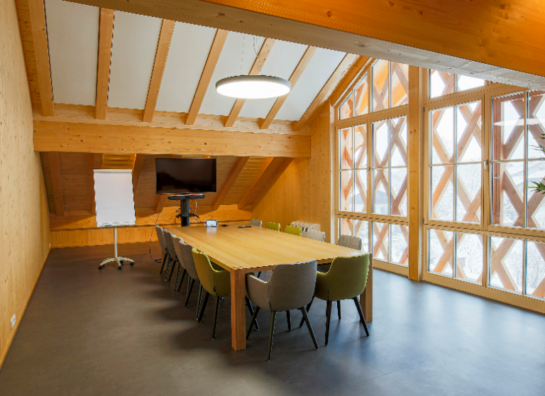 Holztisch im Konferenzraum, bunte Stühle, Monitor, Flipchart, Fenster mit Holzkreuzmustern.