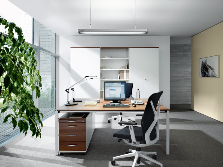 Schreibtisch mit Monitor, Lampe, Stuhl, Telefon, Flasche; helle Fenster.