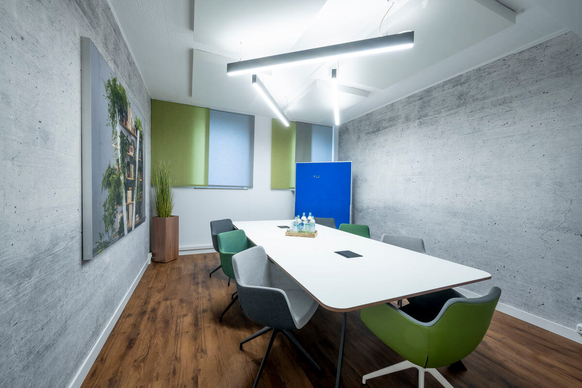 Moderner Meetingraum mit Betonoptik-Wand, Milchglasfenstern in Hellblau und Grün sowie hängender Linearbeleuchtung über einem weißen Konferenztisch mit Stühlen
