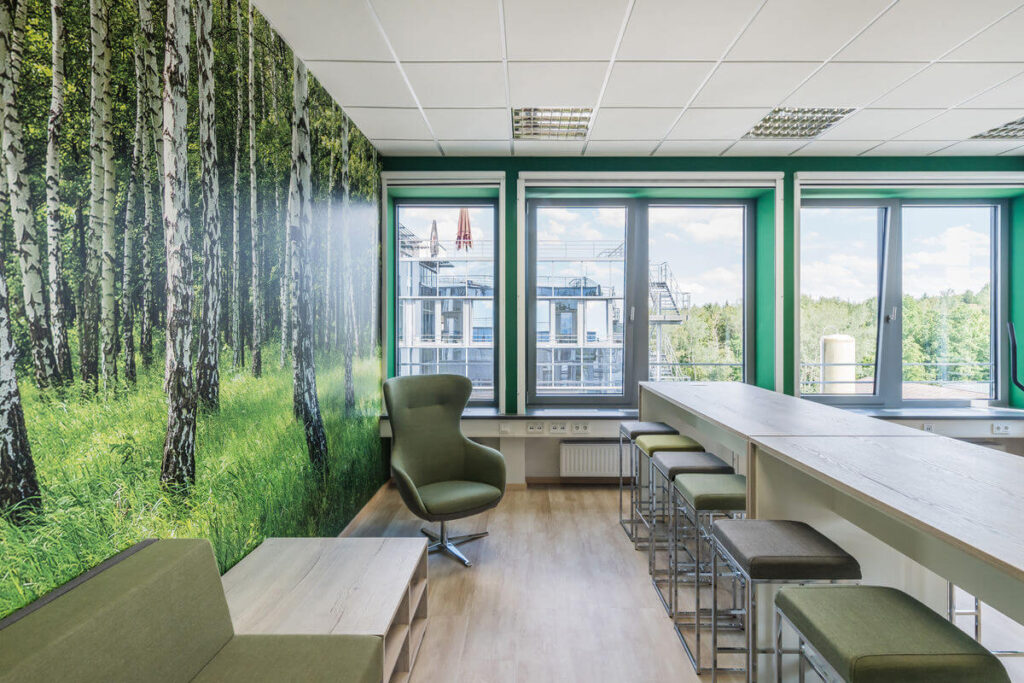 Offener Meetingraum mit Tapete, grün gepolstertem Stuhl, Sitzbank und hellen Arbeitsplätzen mit moderner Deckenbeleuchtung