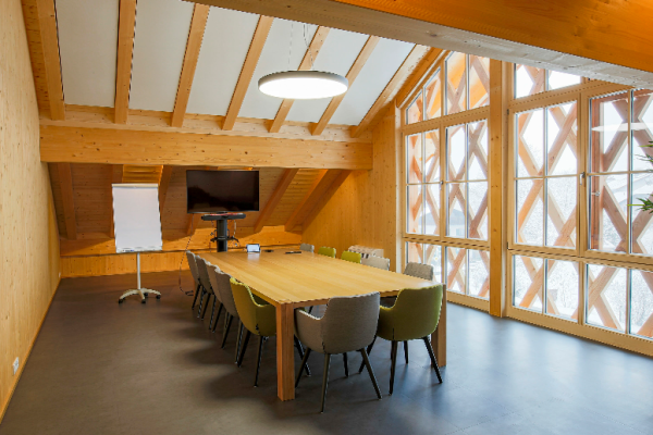 Holztisch im Konferenzraum, bunte Stühle, Monitor, Flipchart, Fenster mit Holzkreuzmustern.