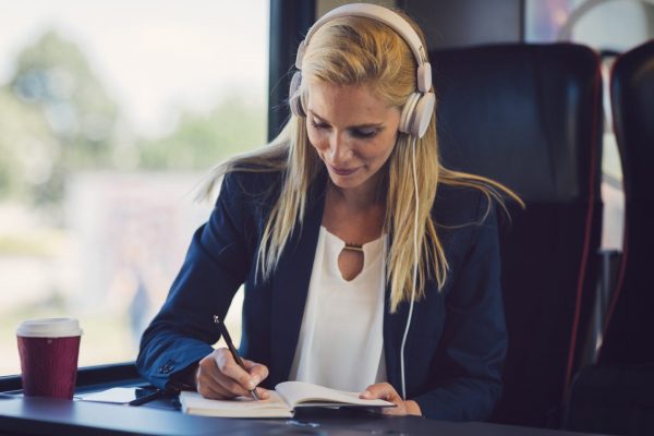 Frau mit Kopfhörern schreibt im Zug, Kaffee neben ihr.