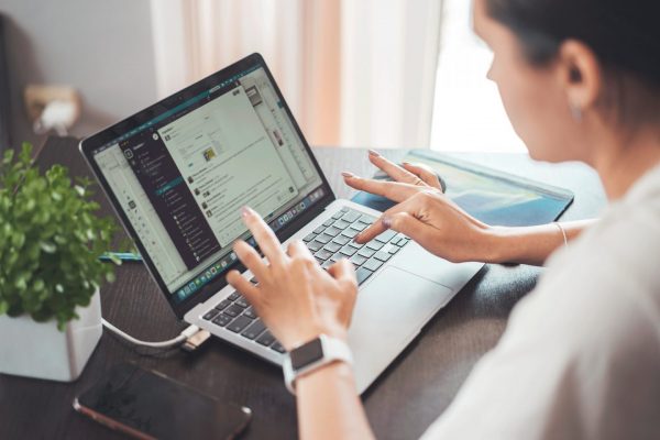 Person am Laptop mit Textnachrichten, Uhr, Pflanze und Smartphone auf Tisch.